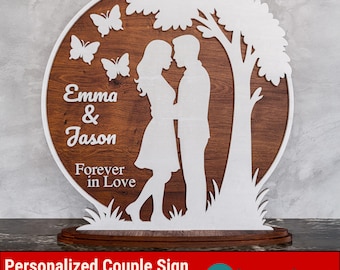 Può includere: Un'insegna in legno con una coppia che si abbraccia sotto un albero, con farfalle e le parole "Emma & Jason" e "Forever in Love". Lo sfondo è in legno marrone e le figure sono ritagliate in bianco.