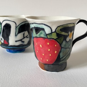 Op de afbeelding: Twee handbeschilderde keramische mokken. Eén mok heeft een zwart-wit abstract ontwerp met een blauwe basis. De andere mok heeft een rood aardbeienontwerp met een zwart handvat en een zwarte en bruine basis. De mokken staan op een witte achtergrond.