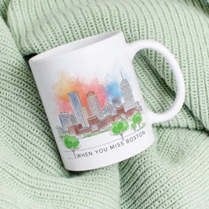 Peut inclure: Mug en céramique blanche avec une illustration de l'horizon de Boston. Le design présente un fond aquarelle coloré et le texte "WHEN YOU MISS BOSTON". La tasse représente des bâtiments emblématiques et des arbres.