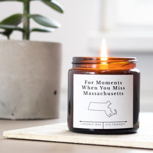 May include: A lit, amber-colored glass jar candle with a white label that reads "For Moments When You Miss Massachusetts" with a Massachusetts outline. The candle is labeled "Coconut Wax" and "Eco-Friendly".