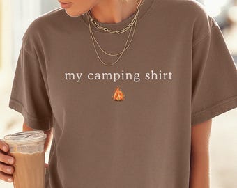 May include: A brown t-shirt with the text "my camping shirt" and a small campfire graphic. The shirt has short sleeves and a relaxed fit. The person in the image is wearing gold jewellery and a black cap.