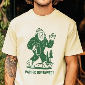 Puede incluir: Camiseta color crema con una ilustración verde de Bigfoot sosteniendo una botella, saludando y caminando cerca de dos pinos. El texto "PACIFIC NORTHWEST" está impreso debajo.
