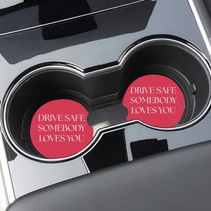 May include: A car interior with two cup holders, each containing a red coaster. The coasters have the white text "DRIVE SAFE SOMEBODY LOVES YOU". The cup holders are set in a silver and black console.