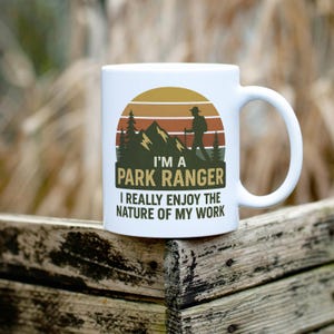 Peut inclure: Mug en céramique blanche avec une anse. Le mug présente un graphisme de paysage de montagne avec un randonneur et le texte "I'M A PARK RANGER I REALLY ENJOY THE NATURE OF MY WORK" en vert et blanc.