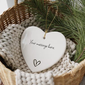 May include: A white heart-shaped ornament with the text "Your message here" and a small heart graphic. The ornament hangs from a gold string and rests in a woven basket with a white knitted blanket and evergreen branches.