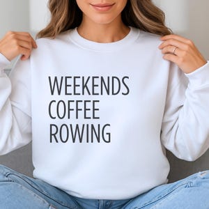 Puede incluir: Sudadera blanca con las palabras "WEEKENDS COFFEE ROWING" impresas en negro. La sudadera es de cuello redondo y la sostiene una persona. La persona lleva vaqueros azules.