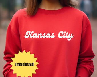 May include: A red sweatshirt with the words "Kansas City" embroidered in white script across the chest. A yellow starburst-shaped tag with the word "Embroidered" is on the lower left.