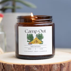 May include: A lit, brown glass jar candle labeled "Camp Out" with a camping scene graphic. The candle is 9 oz and made of 100% natural coconut wax. The candle sits on a wooden surface.