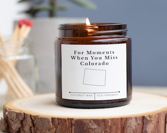 Op de afbeelding: Een brandende bruine glazen potkaars met de tekst "For Moments When You Miss Colorado" en een omtrek van Colorado. De kaars staat op een houten schijf met een ruwe schorsrand. De kaars is gelabeld met "Coconut Wax" en "Eco-Friendly".
