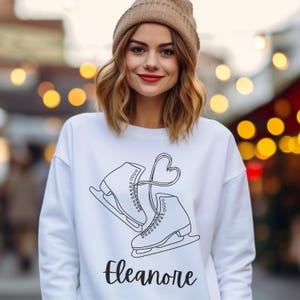 May include: White sweatshirt featuring a black graphic of two ice skates connected by a heart-shaped design. The name "Eleanore" is printed in a cursive font below the skates. The person in the image is wearing a brown beanie.