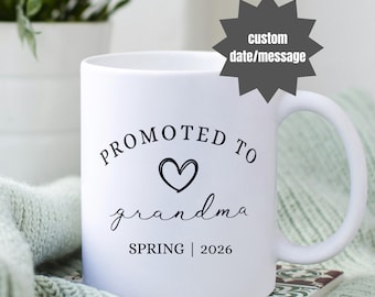 Puede incluir: Taza de cerámica blanca con las palabras "PROMOTED TO grandma" en escritura negra, un símbolo de corazón y "SPRING | 2026". Una estrella personalizada de fecha/mensaje está en la parte superior derecha. La taza está sobre una superficie estampada.