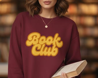 May include: A burgundy sweatshirt with the words "Book Club" in a yellow, retro font. The sweatshirt is being worn by a person holding a book. The background is a blurred bookshelf.