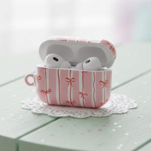 May include: Pink and white striped AirPods case with red bow accents, resembling a teacup. The case is open, revealing white AirPods. The case sits on a white lace doily on a light green surface.