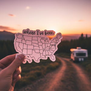 Puede incluir: Una pegatina blanca con la forma de los Estados Unidos, con contornos de estados, sostenida contra una puesta de sol. La pegatina tiene el texto "where I've been" en la parte superior. Una caravana es visible en el fondo en un camino de tierra.