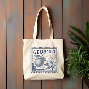Può includere: Borsa tote color crema con un design a timbro blu e bianco con la scritta "GEORGIA". Il timbro include una pesca, un fiore di corniolo e il contorno di un paesaggio. La borsa ha due manici.