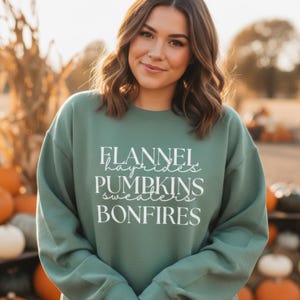 Może przedstawiać: Zielona bluza z białym napisem: "Flannel, hayrides, pumpkins, sweaters, bonfires". Bluza jest w jednolitym zielonym kolorze. Tło jest rozmyte, ale wydaje się być na zewnątrz z dyniami.