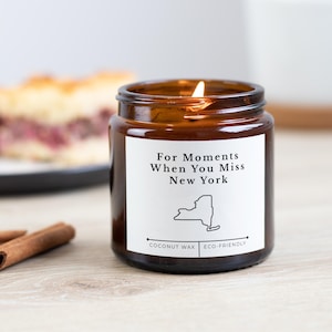 Könnte beinhalten: Ein brennendes bernsteinfarbenes Glas-Kerzenglas mit einem weißen Etikett mit der Aufschrift "For Moments When You Miss New York" mit einer Umrisslinie des Bundesstaates New York. Die Kerze ist mit "Coconut Wax" und "Eco-Friendly" beschriftet. Zimtstangen und ein Stück Kuchen sind im Hintergrund.
