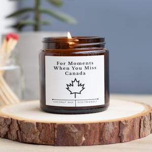 May include: A lit, brown glass jar candle with a white label that reads "For Moments When You Miss Canada" and a maple leaf graphic. The candle sits on a wooden slice. The candle is made with coconut wax and is eco-friendly.