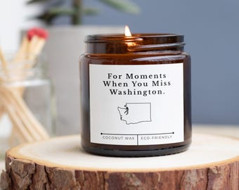 May include: A lit candle in a brown glass jar with a white label. The label reads "For Moments When You Miss Washington." The candle sits on a wooden slice. The candle is made of coconut wax and is eco-friendly.