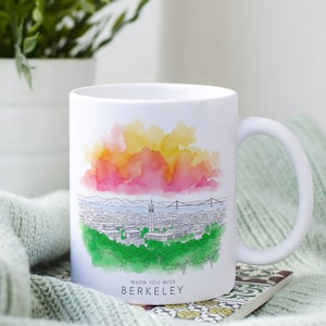May include: White ceramic mug featuring a watercolor design of a cityscape with a bridge and colorful sky. The mug has the text "WHEN YOU MISS BERKELEY" in black. The mug is on a patterned surface.