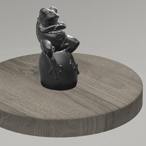 May include: A black frog statue sits atop a round, wooden-textured platform. The frog is detailed with visible eyes and limbs, and the platform has a natural wood grain appearance.