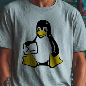 May include: A light grey t-shirt with a black and yellow cartoon penguin holding a computer screen. The penguin has a yellow beak and feet. The t-shirt is a crew neck style.