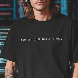 Peut inclure: T-shirt noir avec le texte "You can just build things" en blanc. La personne porte des lunettes de soleil et un casque. Des écrans d'ordinateur avec du code sont en arrière-plan.