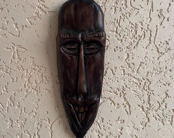 Pode incluir: Uma máscara de madeira entalhada castanha escura com um nariz comprido e um sorriso largo e dentuço. A máscara tem um acabamento liso e polido e está montada numa parede bege texturizada. O design da máscara sugere uma influência tribal africana.