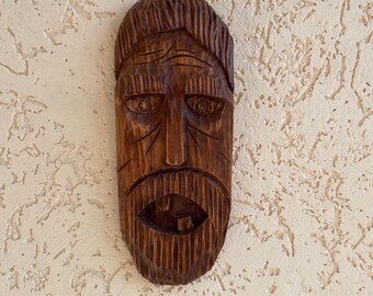 Pode incluir: Uma máscara de madeira esculpida com um rosto detalhado, incluindo barba, bigode e testa franzida. A máscara é de cor castanho médio e parece ser feita à mão. O fundo é uma parede texturizada esbranquiçada.