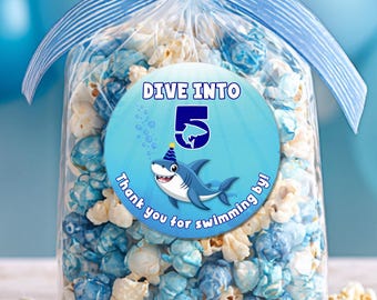 May include: A clear cellophane bag filled with blue and white popcorn, tied with a blue ribbon. A round sticker on the bag reads "DIVE INTO 5" with a cartoon shark. The text "Thank you for swimming by!" is also on the sticker.