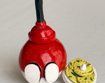 Peut inclure: Une sculpture en céramique fantaisiste faite à la main représentant une figurine ronde rouge et blanche avec une poignée noire, ressemblant à un déboucheur. À côté, une petite assiette blanche contient de petits pierogis jaunes, garnis de crème blanche et d'herbes vertes.