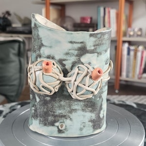 Könnte beinhalten: Eine handgefertigte Keramikvase mit hellblau-grau melierter Glasur. Die Vase hat zwei dekorative, gewebte Tonelemente mit orangefarbenen Akzenten. Die Vase steht auf einem silbernen Metallständer.