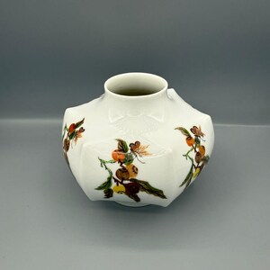 May include: A white ceramic vase with a unique shape and a wide opening. The vase is decorated with colorful floral and fruit designs in shades of orange, brown, and green. The vase has a slightly textured surface.