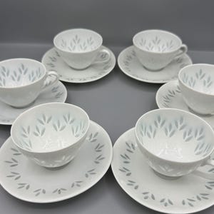 Op de afbeelding: Een set witte kopjes en schotels, elk versierd met een subtiel, lichtblauw bladpatroon. De kopjes hebben een ronde vorm met een oor, en de schotels zijn cirkelvormig. De set is geplaatst op een grijze ondergrond.