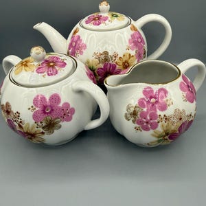 Può includere: Un servizio da tè in porcellana bianca composto da teiera, zuccheriera e lattiera. Ogni pezzo è decorato con motivi floreali rosa, gialli e marroni. Il set ha bordi dorati e un design classico.