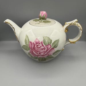 Può includere: Teiera in porcellana bianca decorata con una grande rosa rosa e foglie verdi. La teiera ha un bocciolo di rosa sul coperchio e dettagli dorati sul manico e sul beccuccio. Design classico.
