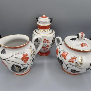 Puede incluir: Un juego de té de porcelana blanca que consta de una jarra de leche, un azucarero con tapa y un recipiente alto con tapa. Cada pieza presenta un patrón pintado de bayas naranjas, hojas negras y detalles dorados con bordes en naranja y oro.