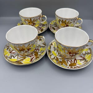May include: Four white teacups with gold trim and matching saucers. The cups and saucers have a yellow and brown floral design. The handles and rims of the cups are gold.