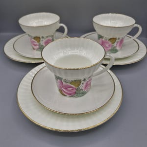 Peut inclure: Un ensemble de trois tasses et soucoupes blanches. Les tasses et les soucoupes ont une bordure dorée et un motif texturé. Les tasses présentent un motif floral avec des roses roses et des feuilles vertes. Les soucoupes sont rondes.