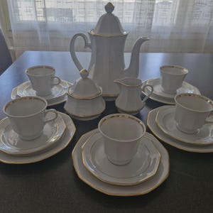 May include: A white porcelain tea set with gold trim. The set includes a coffee pot, sugar bowl, creamer, and six cups with saucers and plates. The pieces are arranged on a dark surface.
