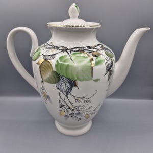 May include: A white porcelain teapot featuring a hand-painted botanical design of green and grey leaves with delicate gold accents. The teapot has a curved handle, a long spout, and a matching lid with a small finial knob on top.