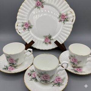 Puede incluir: Un juego de té de porcelana blanca con diseños florales de rosas rosas. El juego incluye un plato grande, tres tazas de té y platillos. Los bordes son festoneados y adornados con oro. Un juego de té clásico y elegante.