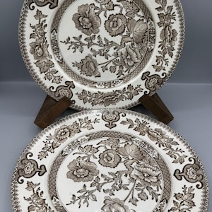 Peut inclure: Deux assiettes en céramique blanche avec un motif floral marron. Les assiettes ont une bordure détaillée et un motif floral central. Elles sont présentées sur un support en bois sur fond gris.