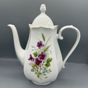 May include: White porcelain teapot with a floral design featuring purple pansies, green leaves, and small white and blue flowers. The teapot has a curved spout, a handle, and a lid with a decorative finial. Gold trim accents the rim.
