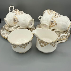 Könnte beinhalten: Ein weißes Porzellan-Teeservice mit goldenen floralen Details. Das Set beinhaltet Teetassen, Untertassen und einen Sahnekännchen. Die Teetassen und der Sahnekännchen haben gebogene Griffe. Die Untertassen und Tassen haben einen goldenen Rand.