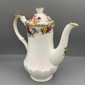 Op de afbeelding: Witte keramische koffiepot met een bloemmotief van roze, gele en rode rozen. De pot heeft een goudkleurige handgreep en rand. Het deksel is ook versierd met hetzelfde bloemmotief.