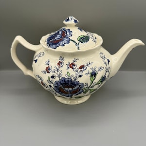 May include: Cream-colored ceramic teapot with a lid, featuring a floral pattern in blue, red, and green. The teapot has a curved spout and handle, and the lid is topped with a small finial. The design is a classic, vintage style.