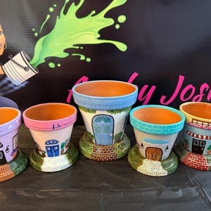 May include: Five terracotta flower pots, each hand-painted to resemble a whimsical house. The pots feature colorful details like doors, windows, and brickwork, with a green base. The background includes a cartoon illustration and the text "by Josie".