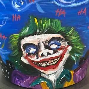 May include: A hand-painted blue ceramic pot featuring a portrait of the Joker with green hair, white face, and a red smile. The background has a starry night design with the word "HA" repeated in red. The pot has a dark rim.