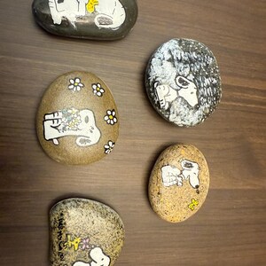 Puede incluir: Cinco piedras pintadas con Snoopy de la tira c&oacute;mica Peanuts. Las piedras son de forma ovalada y est&aacute;n pintadas con detalles blancos y amarillos sobre un fondo oscuro. Algunas piedras tienen a Snoopy con flores.